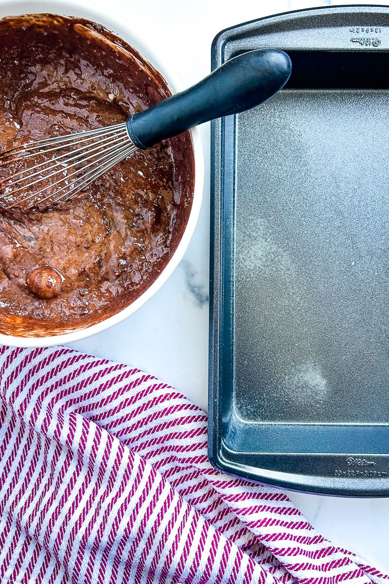 A bowl of cake batter with a whisk and an empty baking pan.