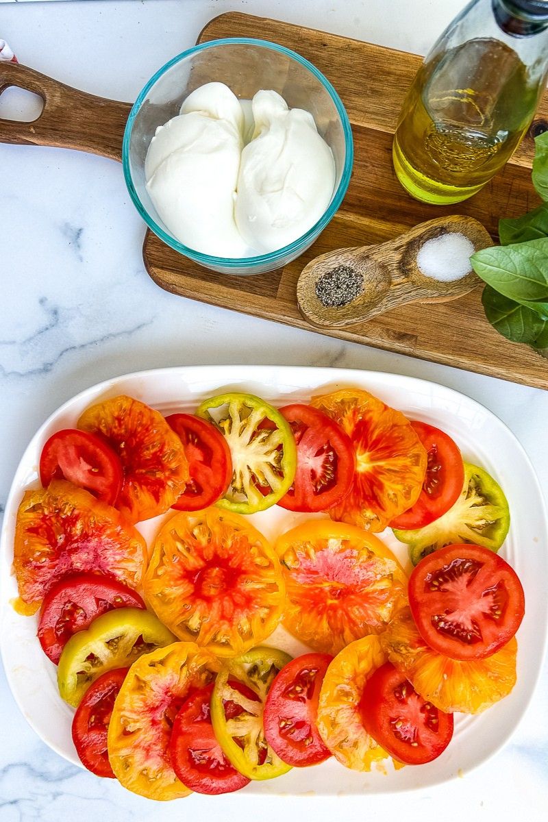 Sliced tomatoes on a white serving plate with a platter of raw ingredients.