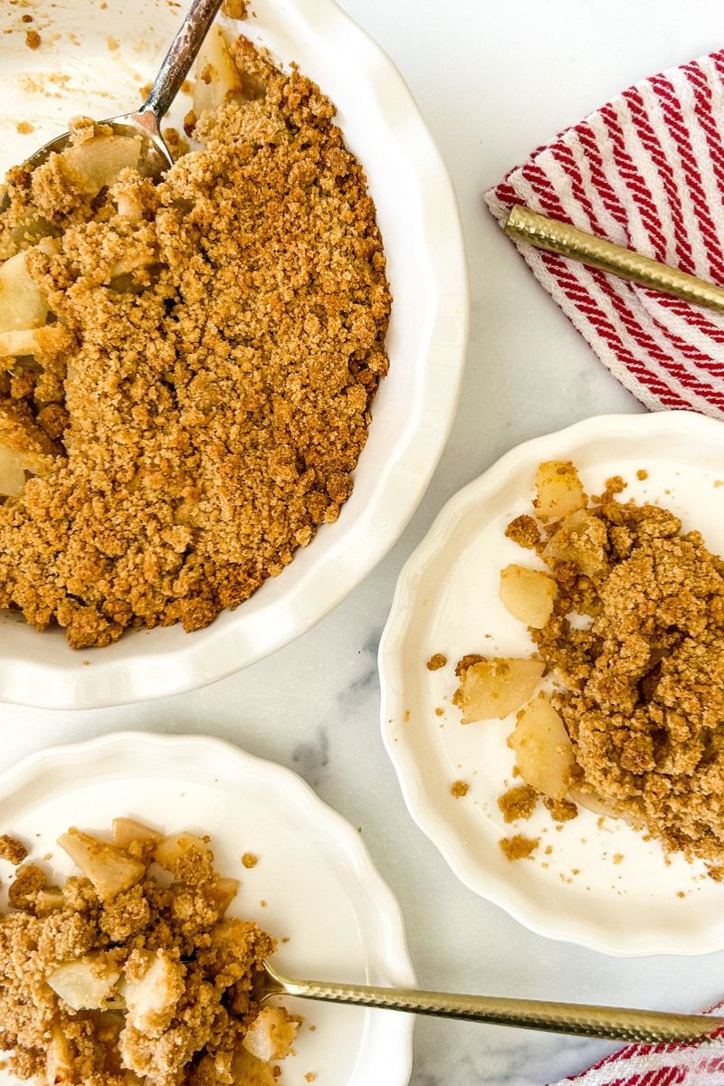 Two plates of pear crumble topped with cream and a pan of pear crumble.