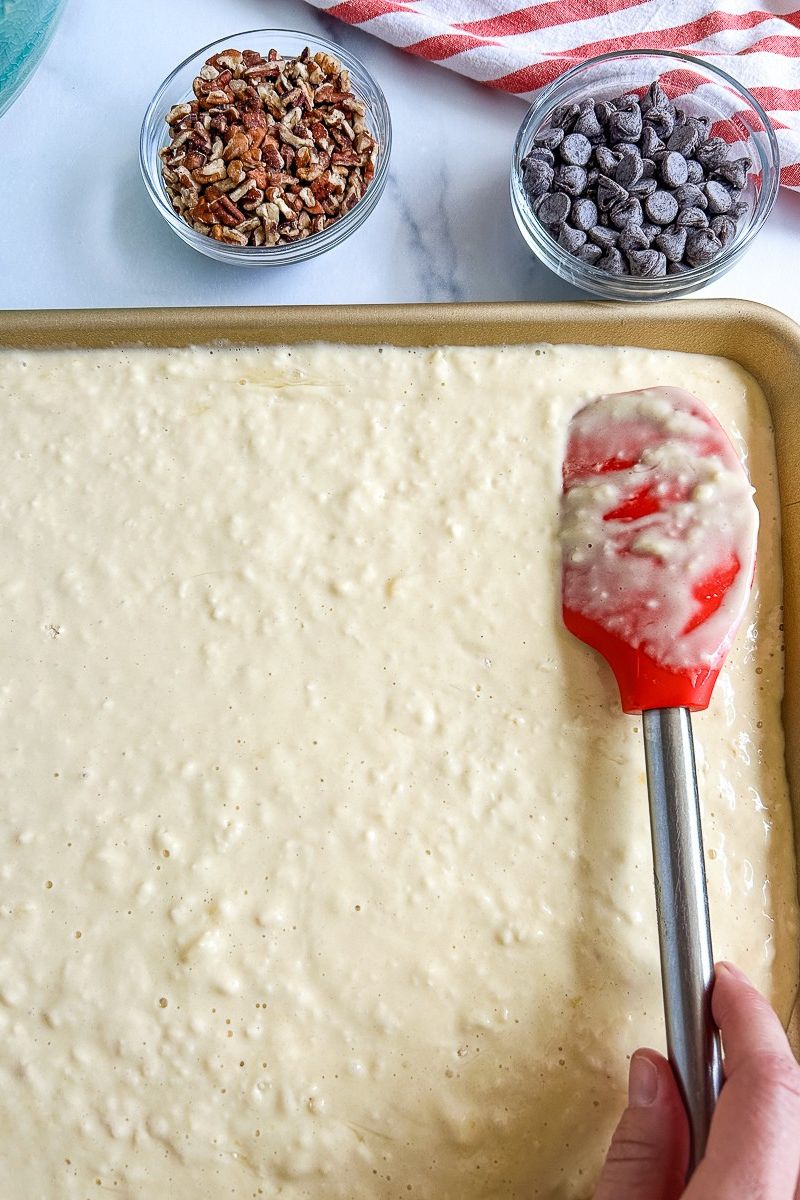A batter of pancake in a sheet pan with rubber scraper and 2 bowls of toppings.