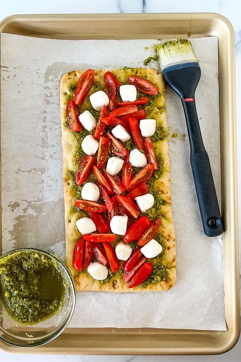 A flatbread on a baking sheet topped with cheese, tomatoes, and pesto sauce.