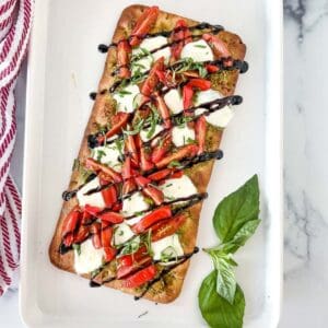 Caprese flatbread with garnish on a white platter and basil leaves.