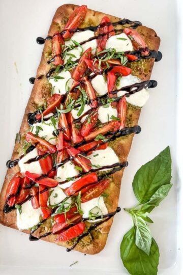 Caprese flatbread with garnish on a white platter and basil leaves.