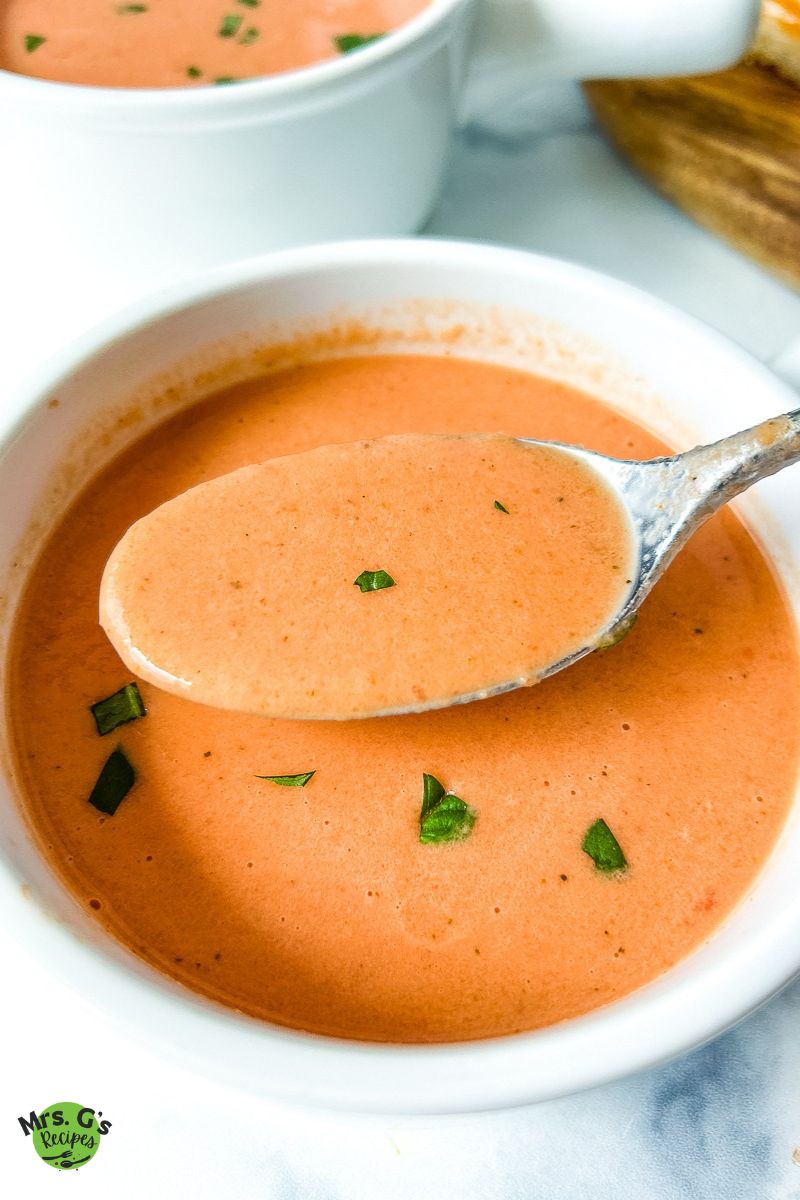 A silver spoon scoops a spoonful of creamy tomato soup from a white bowl. The soup is garnished with small chopped basil pieces.
