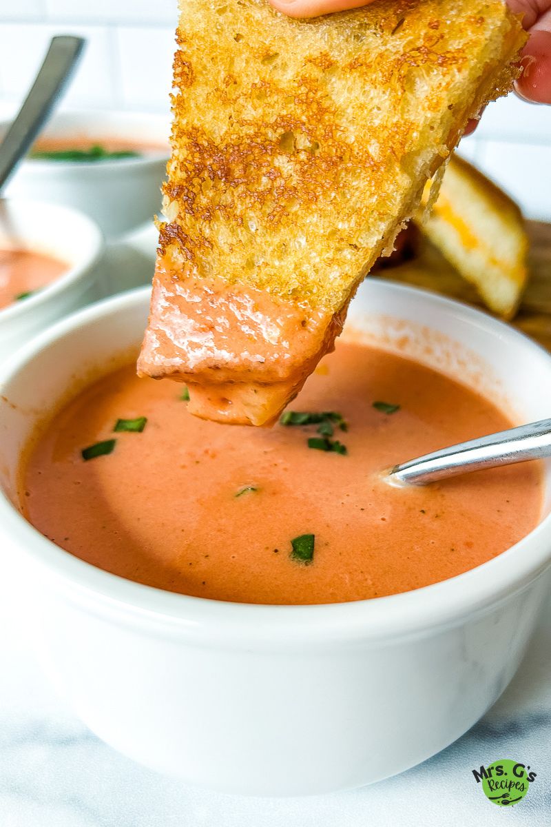 A grilled cheese sandwich half is being dipped into a white bowl of creamy tomato soup. The soup has a garnish of small chopped basil pieces.