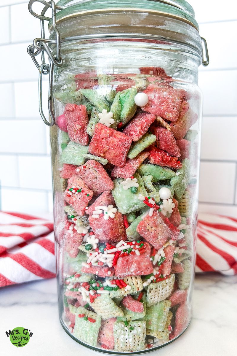 A large glass jar with a latch lid is filled with the finished Christmas muddy buddy snack mix.