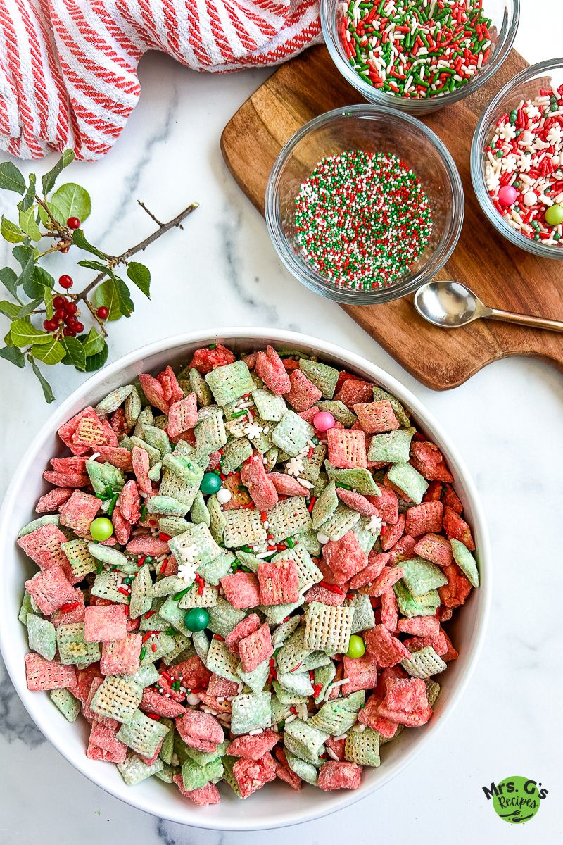 An overhead shot of a large white bowl filled with finished Christmas muddy buddies (red and green Chex cereal coated in powdered sugar and mixed with various Christmas sprinkles). Several smaller bowls of sprinkles are in the background.