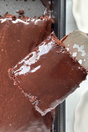A glossy slice of chocolate cake with rich chocolate frosting is being lifted out of a baking pan by a silver serving spatula. The rest of the cake is visible in the pan.