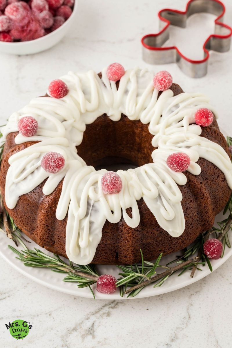 A gingerbread Bundt cake, beautifully decorated with white icing and sugared cranberries, sits on a white plate garnished with fresh rosemary.