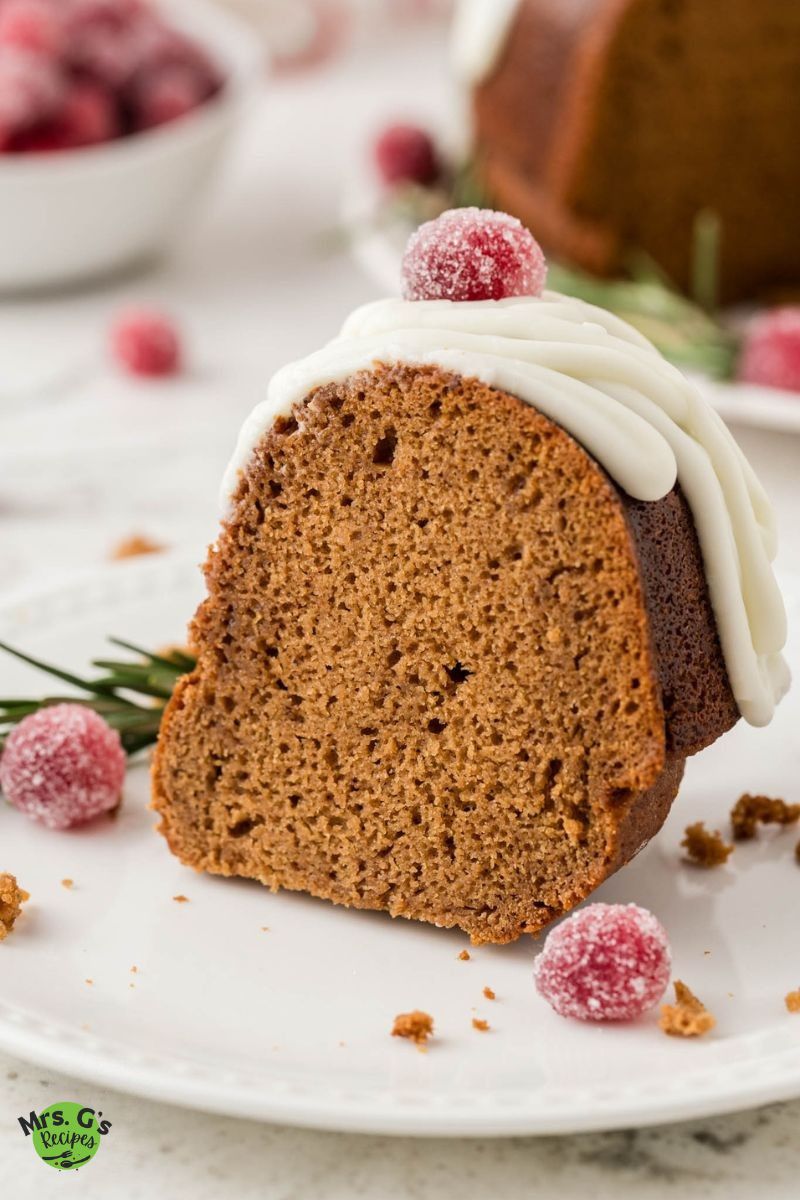 A close-up, eye-level shot of a single slice of gingerbread Bundt cake on a white plate. The slice is topped with white icing and a sugared cranberry.