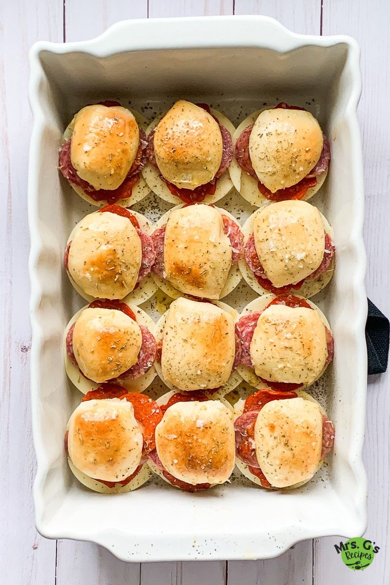 An overhead shot of a white rectangular baking dish with 12 Italian sub sliders. Each slider is topped with a bun brushed with butter and herbs.