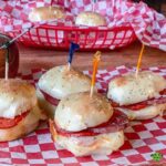 A close-up, eye-level shot of four Italian sub sliders on a red-and-white checkered paper. A red basket with more sliders is in the soft-focus background.