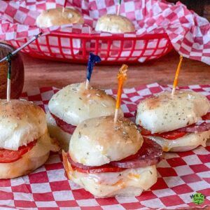 A close-up, eye-level shot of four Italian sub sliders on a red-and-white checkered paper. A red basket with more sliders is in the soft-focus background.