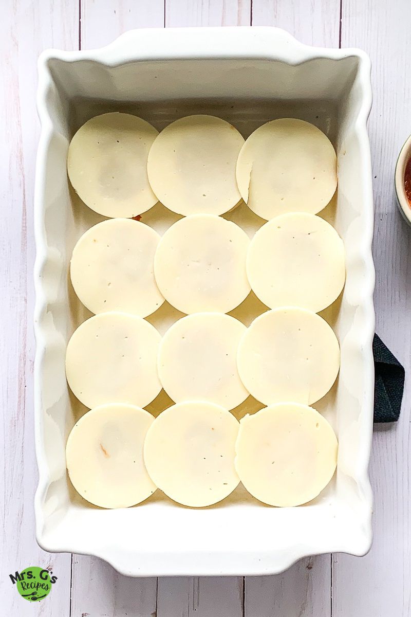 An overhead shot of a white rectangular baking dish filled with the bottom halves of 12 dinner rolls, each topped with a large slice of cheese.