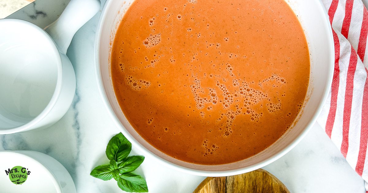 A large white bowl filled with creamy tomato soup sits on a marble surface. A fresh sprig of basil is a garnish. Two smaller white bowls and a wooden board are on the left and right.