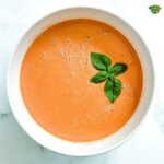 A close-up, overhead shot of a large white bowl filled with creamy tomato soup. A fresh sprig of basil floats in the center of the soup.