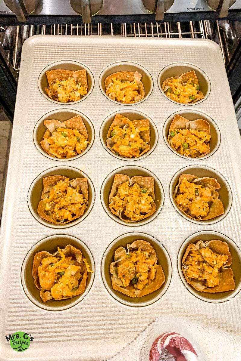 A muffin tin filled with wonton cups, each filled with a mixture of buffalo chicken, cheese, and green onions, is being placed into an oven.