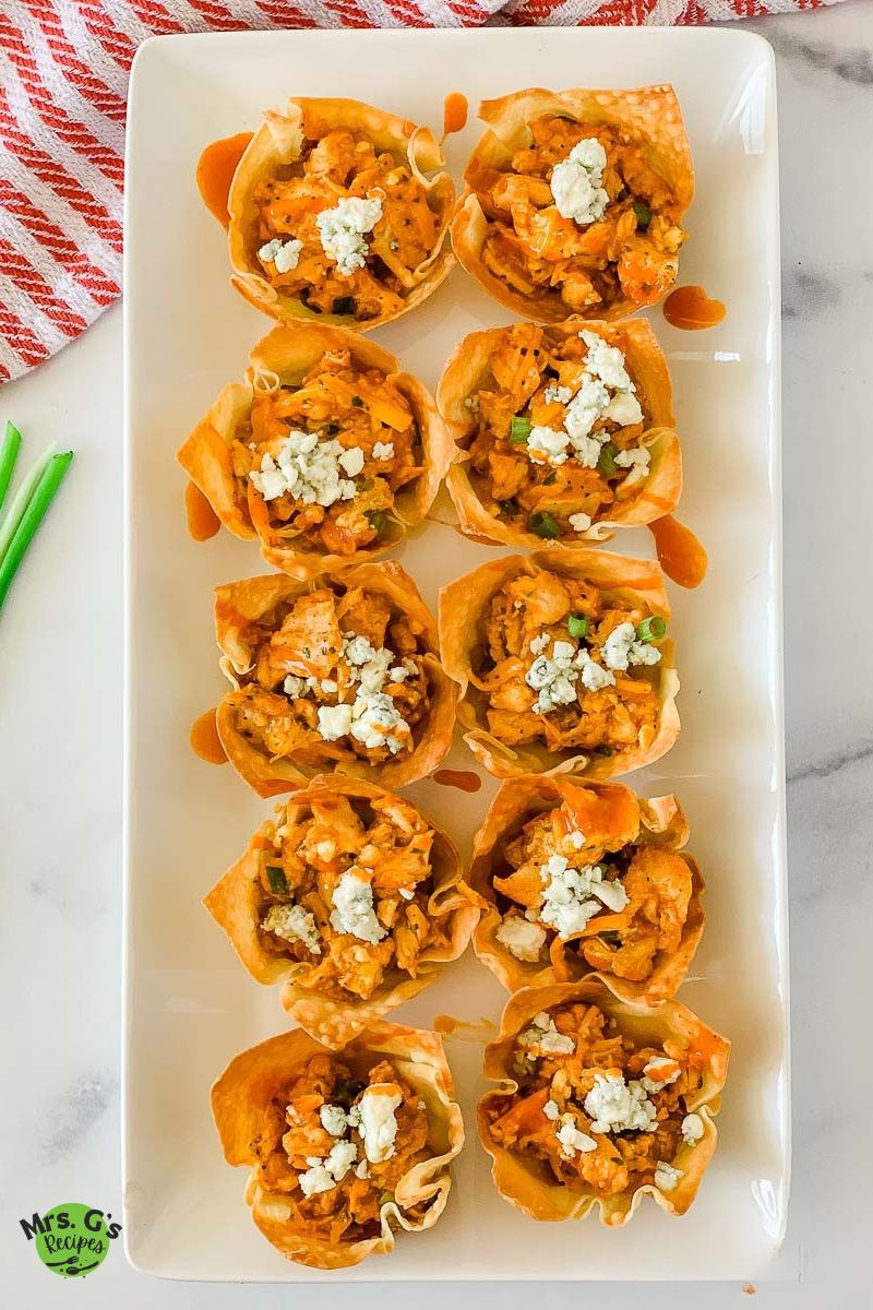 A white rectangular platter holds eight buffalo chicken wonton cups in two rows, each topped with crumbled blue cheese.