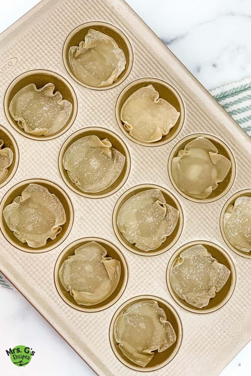 An overhead shot of a muffin tin with uncooked wonton wrappers pressed into each cup, ready to be filled.