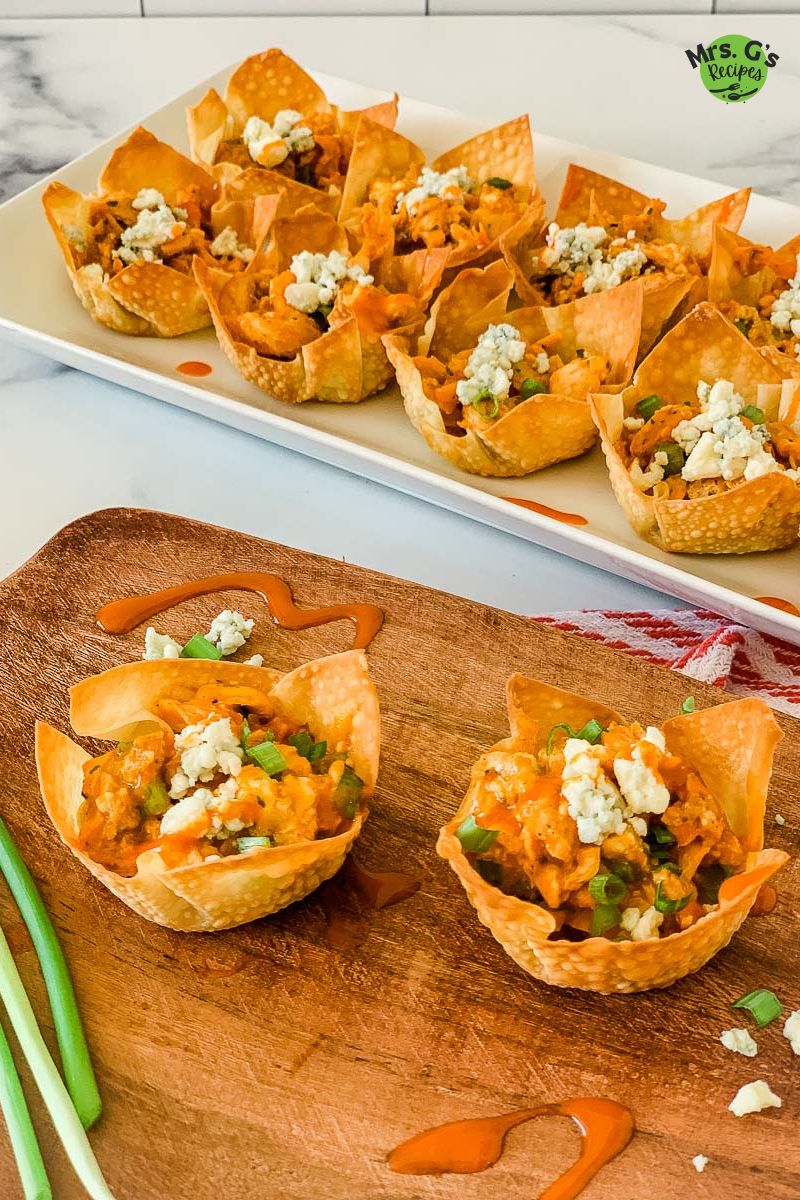 A white rectangular platter holds a dozen buffalo chicken wontons. Two additional wonton cups sit on a wooden cutting board in the foreground.