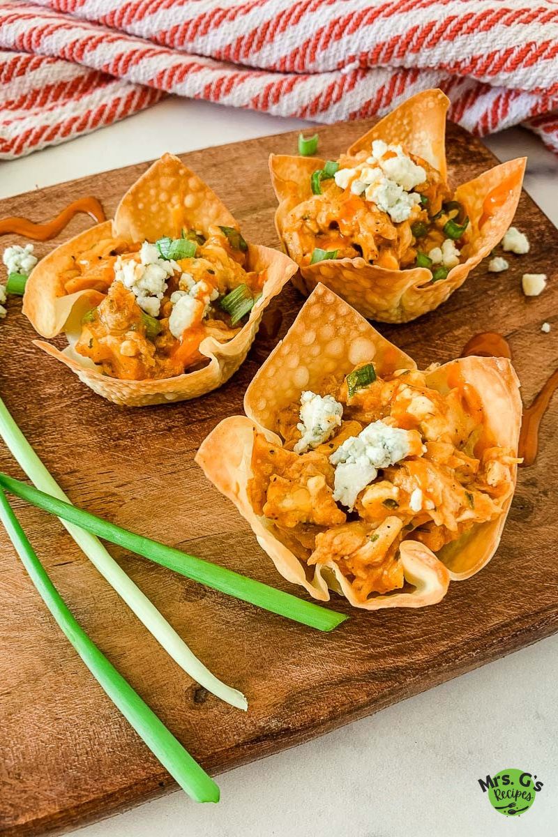 Three individual buffalo chicken wonton cups sit on a wooden cutting board, garnished with blue cheese crumbles and chopped green onions. Drizzles of buffalo sauce and three whole green onions are also visible.