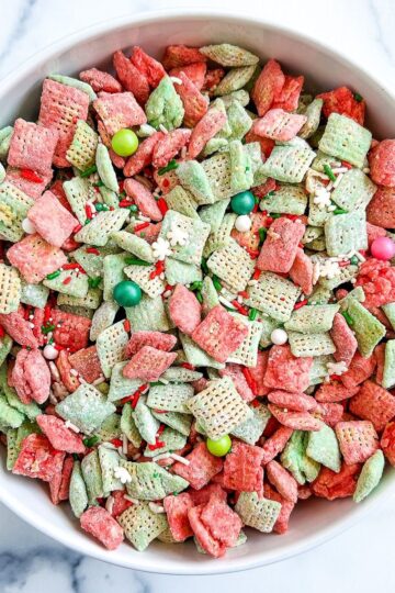 A close-up, overhead shot of a white bowl filled with red and green Chex muddy buddies, featuring various festive sprinkles.