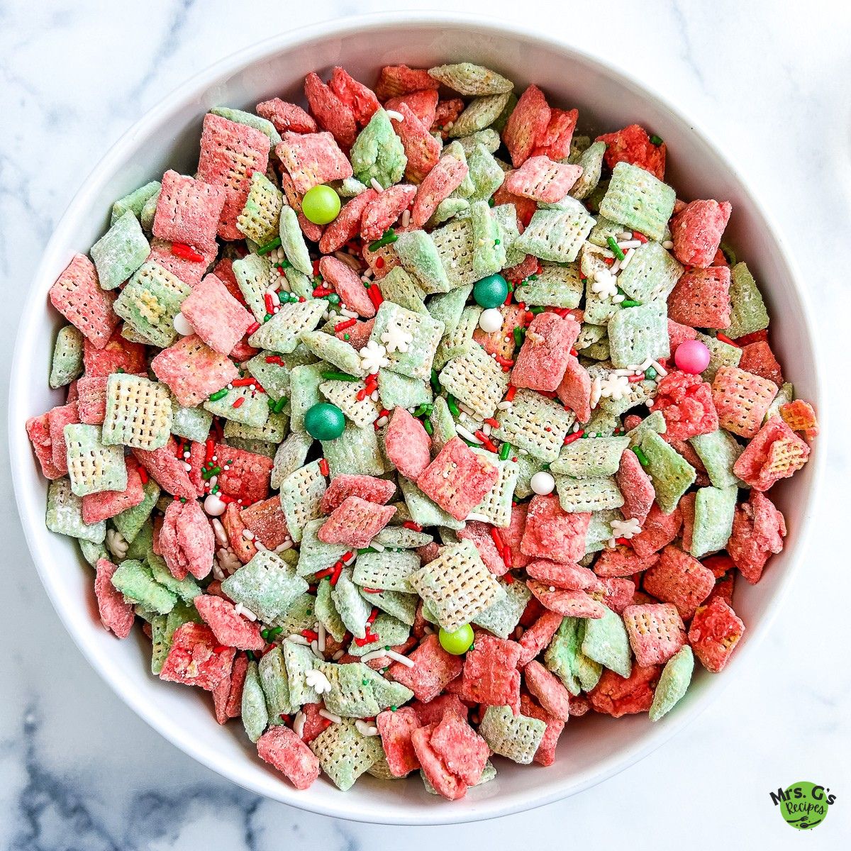 A close-up, overhead shot of a white bowl filled with red and green Chex muddy buddies, featuring various festive sprinkles.