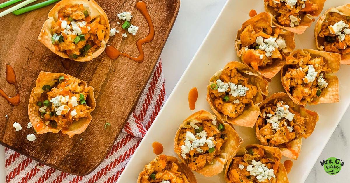 A wide, top-down view showing a wooden cutting board with two buffalo chicken wonton cups next to a white rectangular platter holding a dozen more.