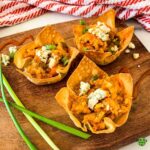 A close-up, overhead shot of three buffalo chicken wonton cups on a wooden cutting board. They are garnished with blue cheese and green onions, and there are sauce drizzles on the board.