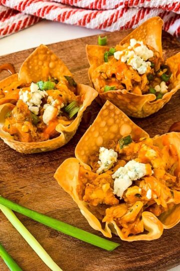 A close-up, overhead shot of three buffalo chicken wonton cups on a wooden cutting board. They are garnished with blue cheese and green onions, and there are sauce drizzles on the board.