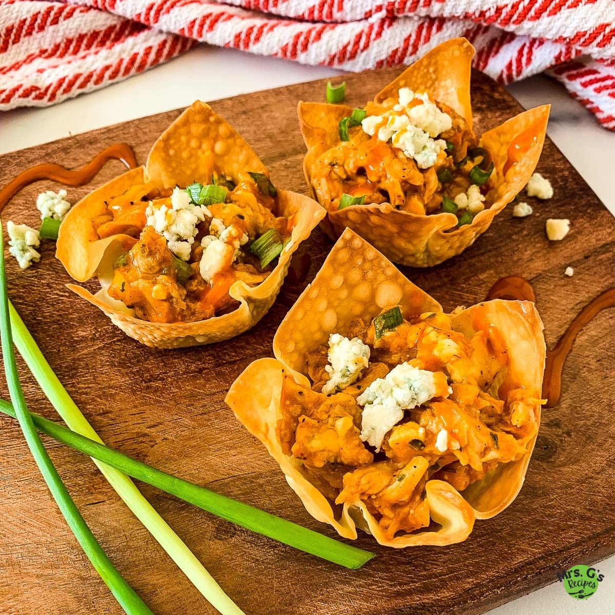 A close-up, overhead shot of three buffalo chicken wonton cups on a wooden cutting board. They are garnished with blue cheese and green onions, and there are sauce drizzles on the board.