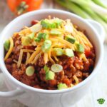 A bowl of chili with cheese and garnish on top.