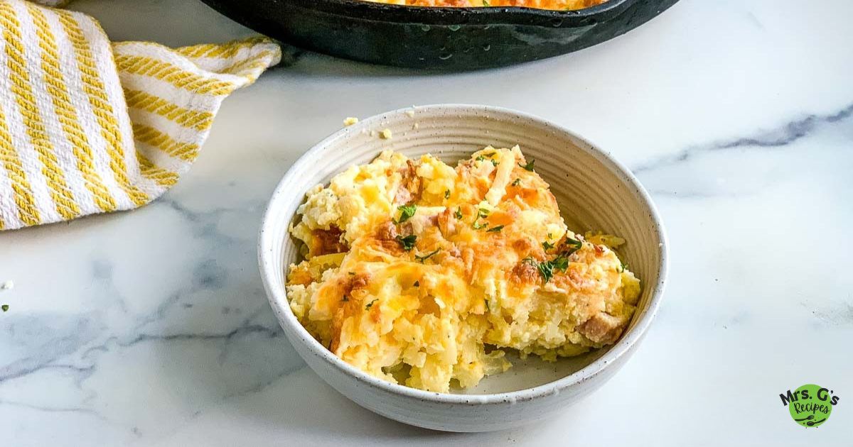 A wide, eye-level shot of a serving of baked ham and hash brown casserole in a small white bowl. The cast iron skillet is visible in the background.