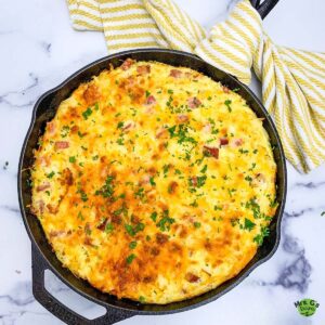 An overhead shot of a baked ham and hash brown casserole in a black cast iron skillet, garnished with fresh parsley. The cheese topping is a golden brown.
