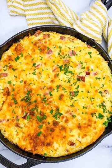 An overhead shot of a baked ham and hash brown casserole in a black cast iron skillet, garnished with fresh parsley. The cheese topping is a golden brown.