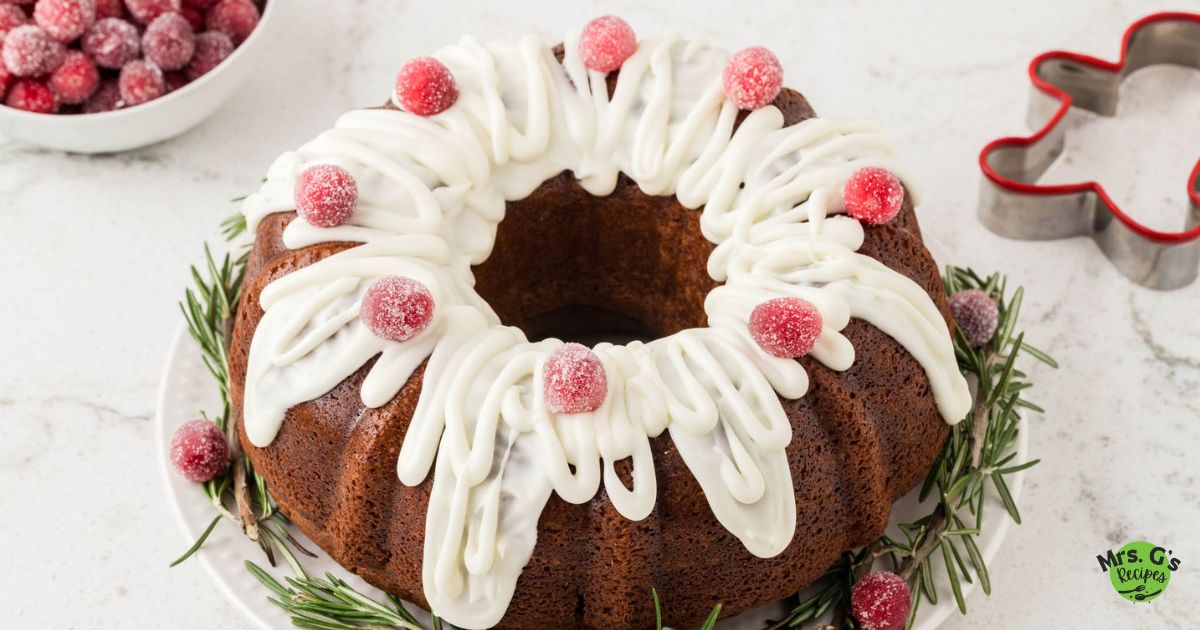A wide, eye-level shot of a beautifully decorated gingerbread Bundt cake on a white plate. It is drizzled with white icing and topped with sugared cranberries, with fresh rosemary sprigs around the base.