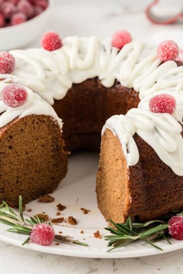 Gingerbread Bundt cake with a slice cut out. The cake is decorated with a icing drizzle and sugared cranberries, and garnished with fresh rosemary sprigs.