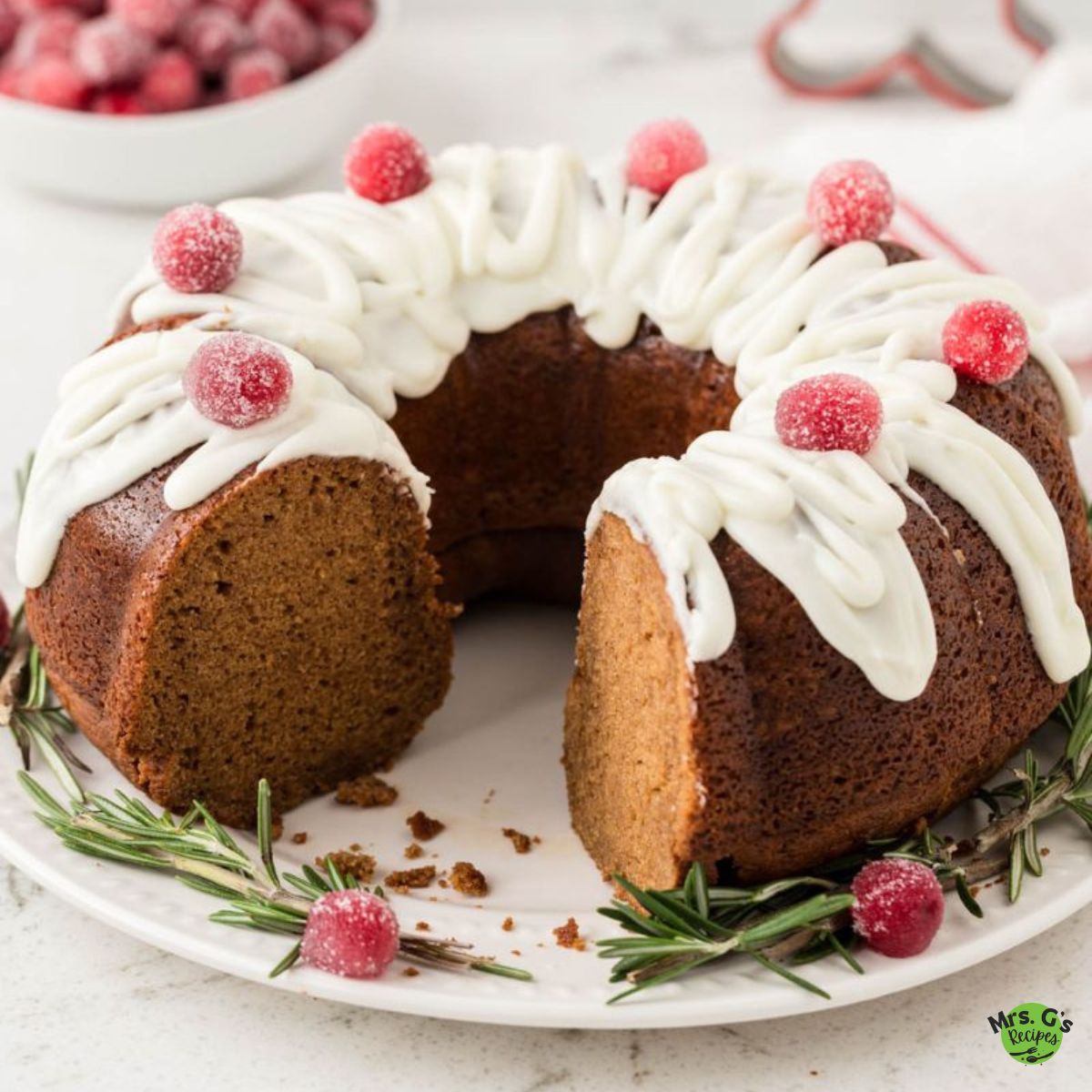 Gingerbread Bundt cake with a slice cut out. The cake is decorated with a icing drizzle and sugared cranberries, and garnished with fresh rosemary sprigs.