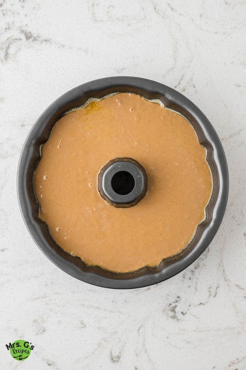 A gray Bundt cake pan sits on a white marble counter, filled with a smooth, light brown gingerbread cake batter.