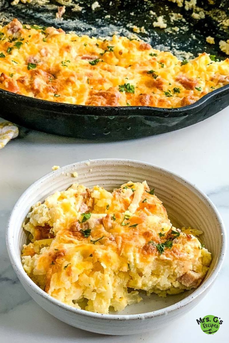 A large cast iron skillet filled with a baked ham and hash brown casserole is in the background, with a serving of the casserole in a white bowl in the foreground.