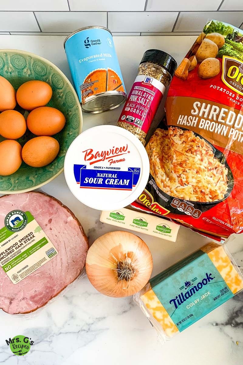 An overhead shot showing the ingredients for a ham breakfast casserole, including a bowl of eggs, a can of evaporated milk, seasonings, a bag of shredded hash brown potatoes, a container of sour cream, a ham steak, an onion, and a block of Colby Jack cheese.