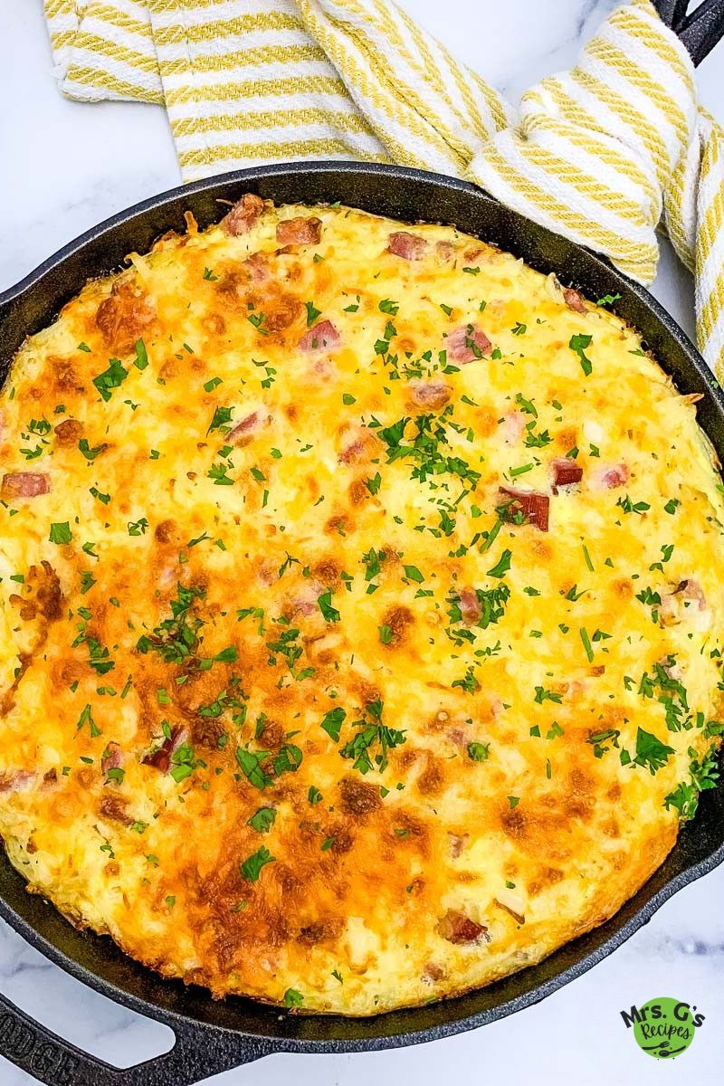 A baked ham and hash brown casserole is shown in a black cast iron skillet, with a yellow and white striped kitchen towel draped over the handle.
