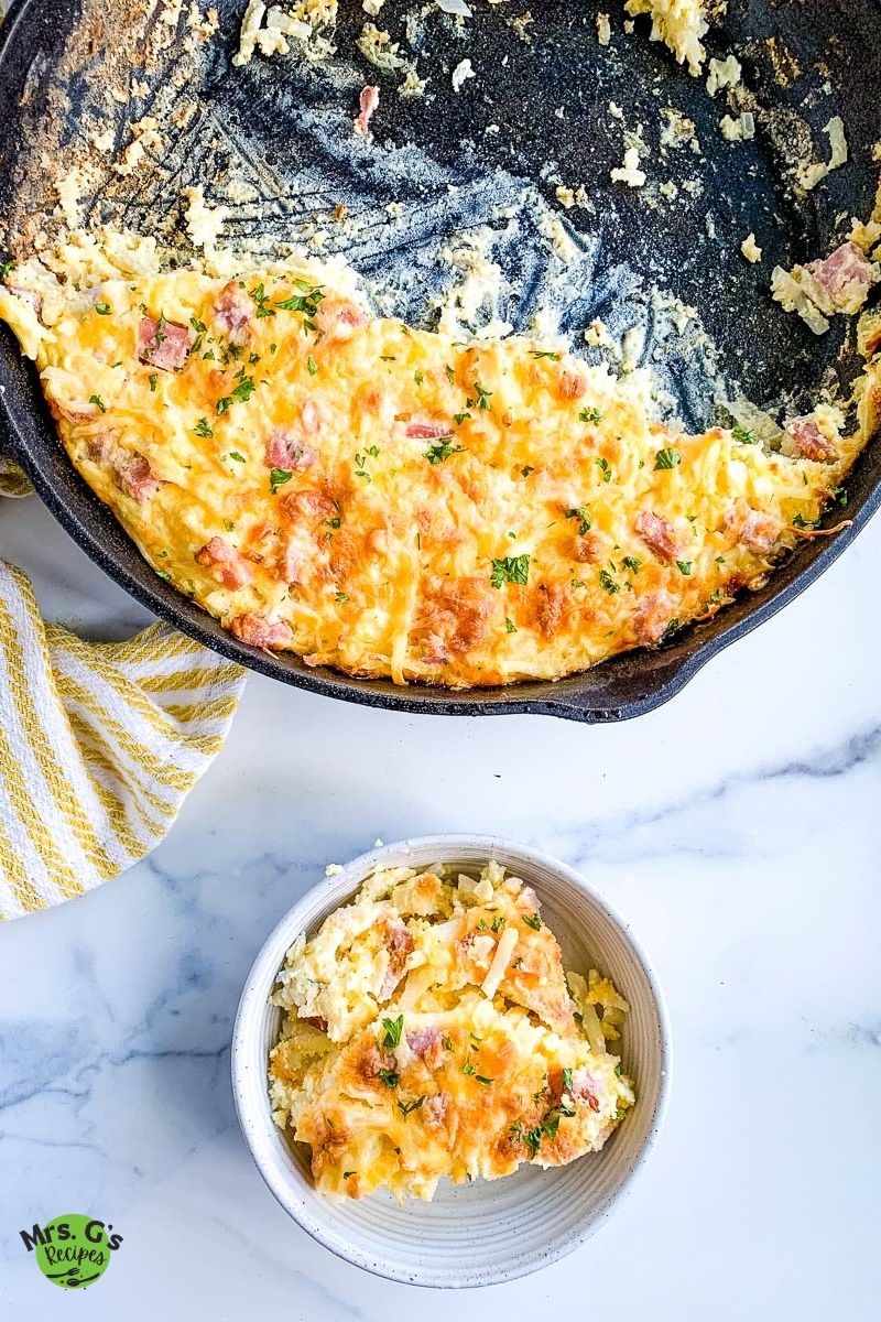 A serving of a baked ham and hash brown casserole sits in a bowl on a marble counter, with the cast iron skillet in the background showing where the serving was removed.