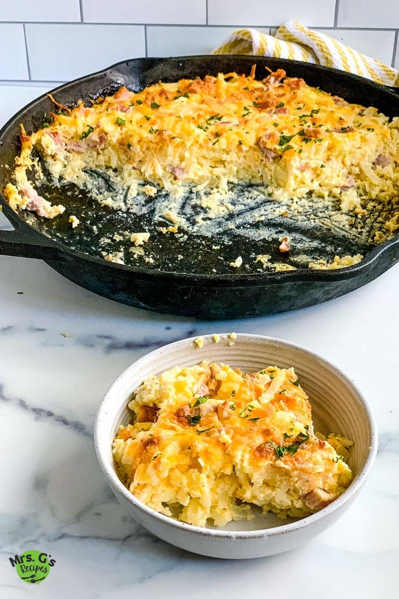 A large cast iron skillet with a baked ham and hash brown casserole sits on a marble surface. A large portion has been scooped out, and a serving is in a bowl in the foreground.