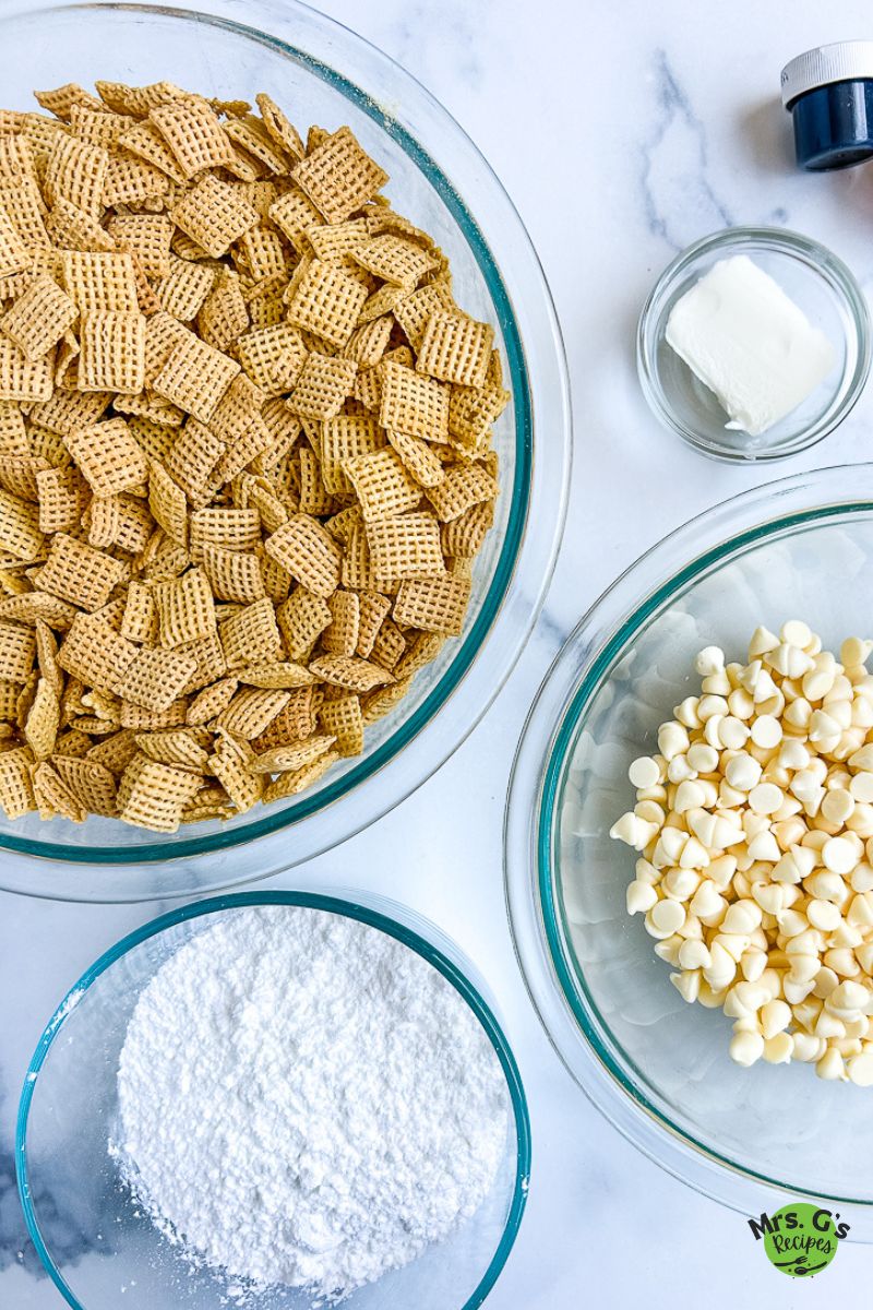 An overhead shot showing the ingredients for muddy buddies on a marble countertop: a large glass bowl of Chex cereal, a bowl of powdered sugar, and a bowl of white chocolate chips.