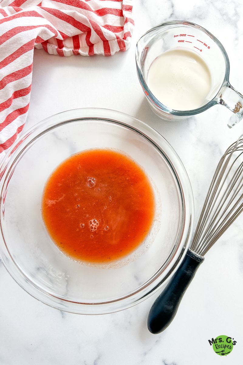 A glass bowl holds a thin, reddish-orange liquid. A whisk with a black handle is next to the bowl, and a measuring cup filled with white liquid is in the background.