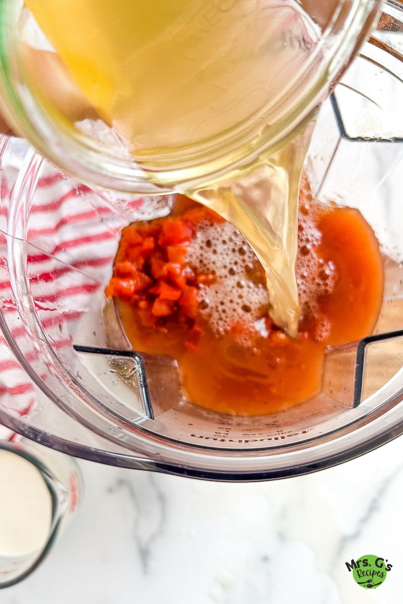 A person pours a light-colored liquid, likely vegetable broth, into a blender filled with diced and crushed tomatoes.