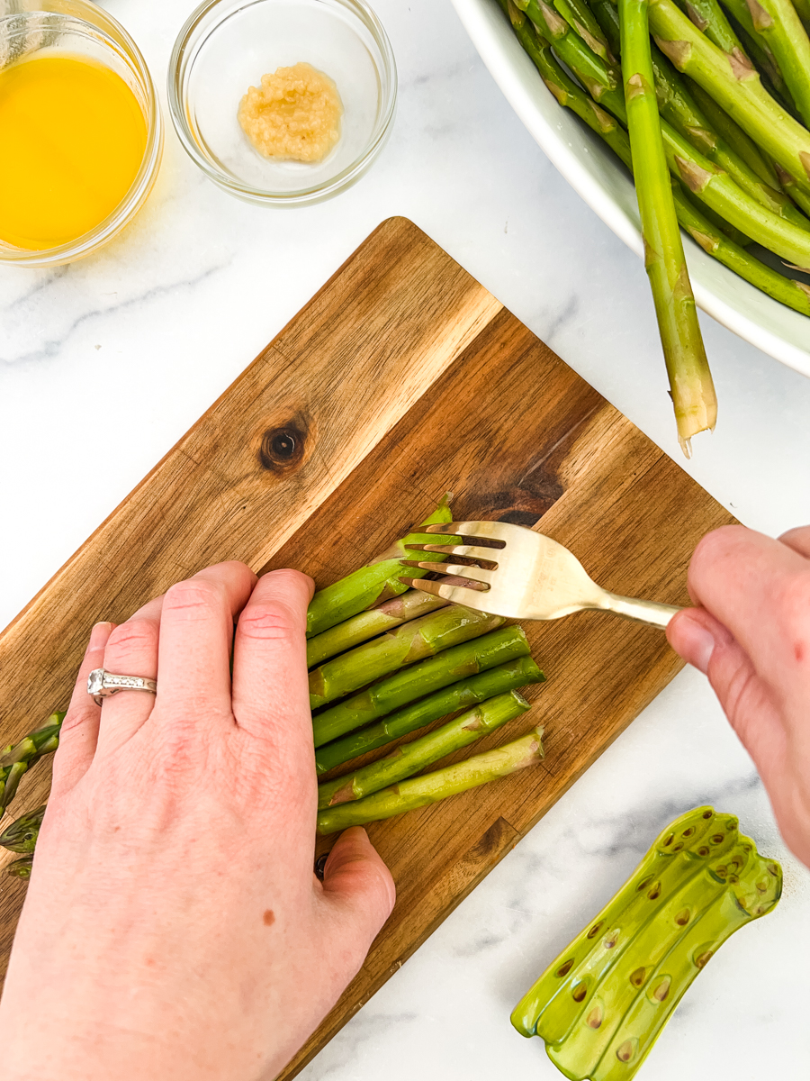 A cutting board with asparagus being poked with a fork. In the background is melted butter, garlic and more asparagus.
