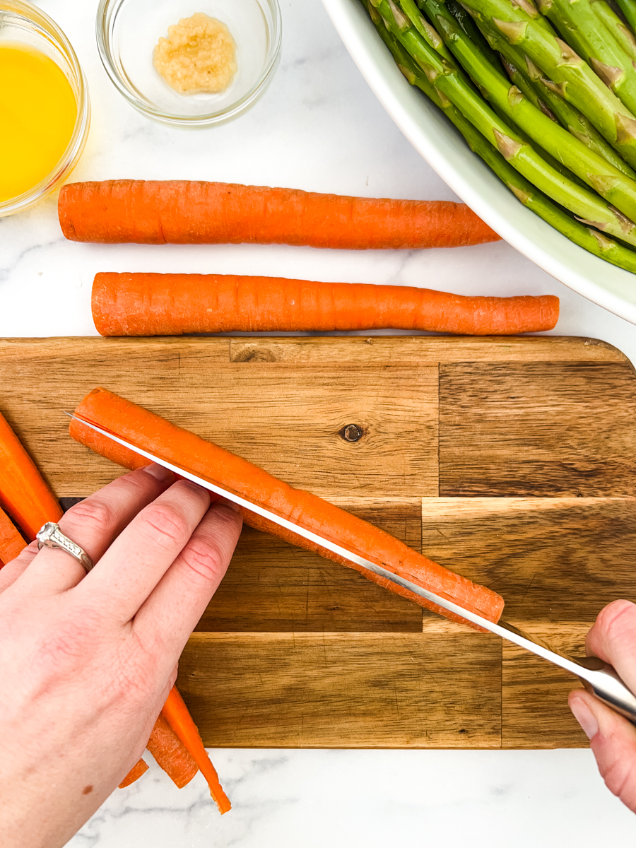A cutting board with a carrot being sliced in half. In the background is melted butter, garlic and asparagus.