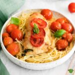 Square format close-up of a serving of Chicken Margherita on a bed of spaghetti, surrounded by red cherry tomatoes and green basil leaves. The chicken breast is topped with sliced tomatoes and cheese.